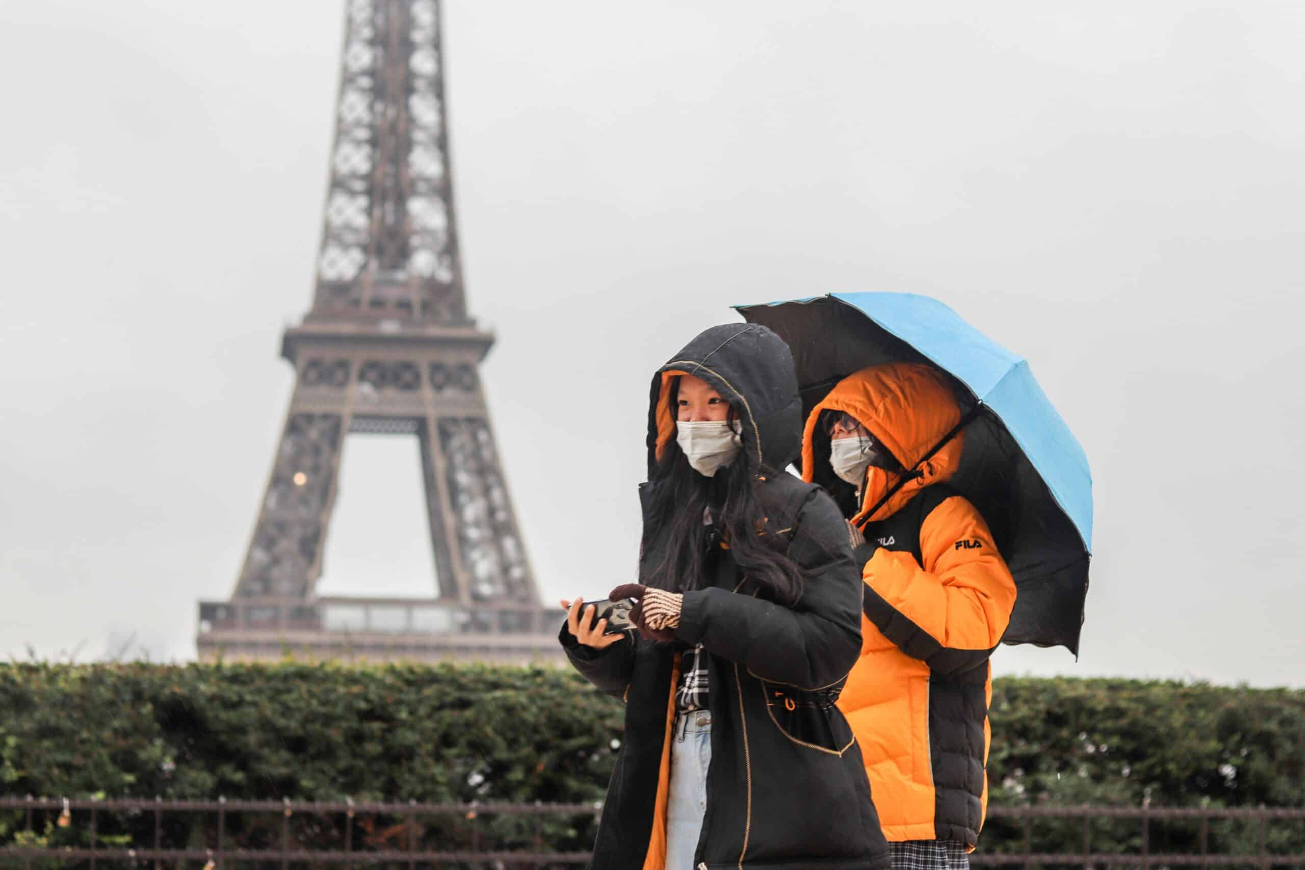 Two people walk in front of the Eiffel Tower in the rain, wearing rain jackets and holding an umberella