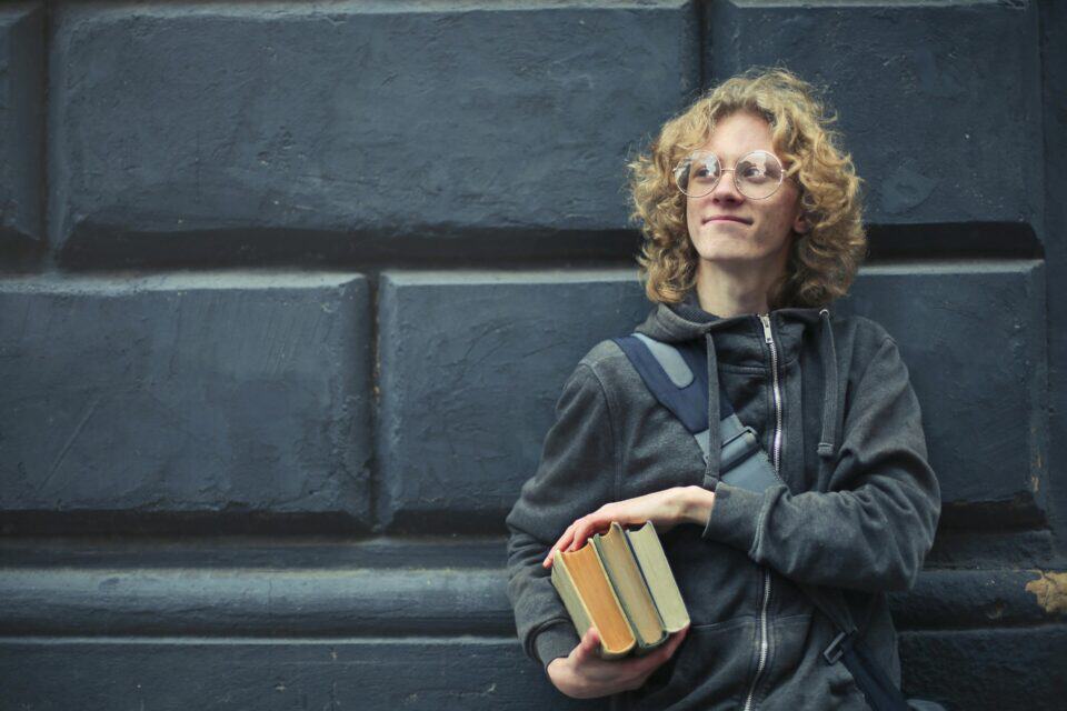 A student stands against a dark wall holding books and smiling