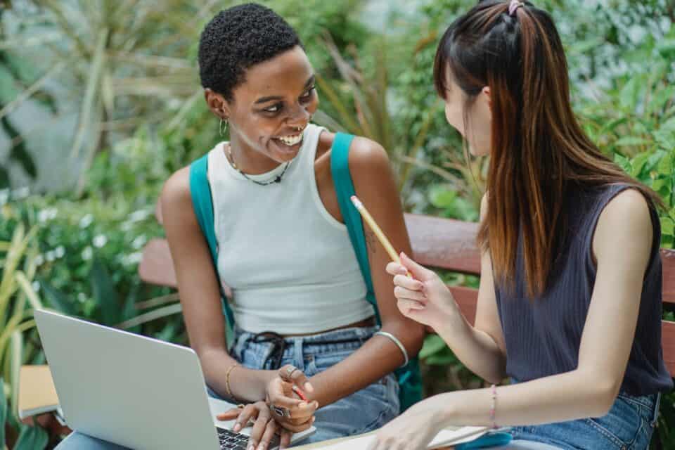 Two female students chatting on park bench with laptop.