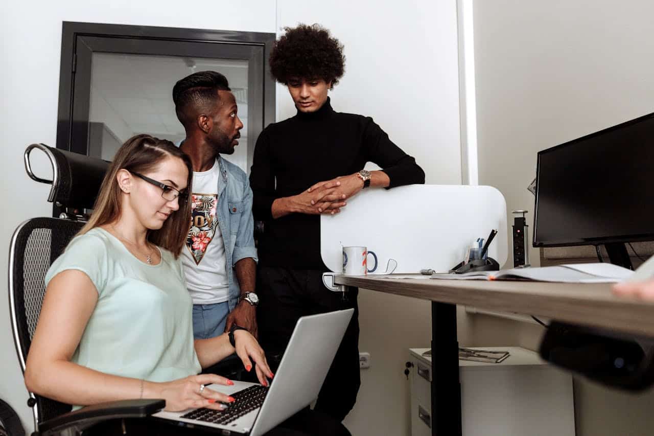 Woman working on laptop with two men chatting behind her.