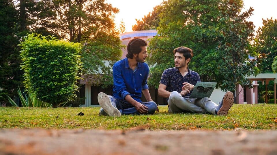 Two male university students sitting on university lawn with a laptop.